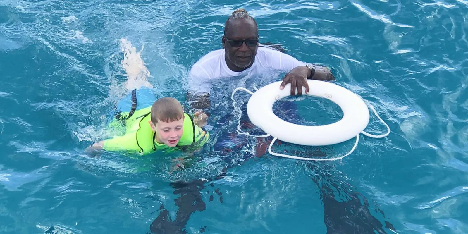 A crew member steadying a life ring beside a young boy swimming in a buoyancy vest