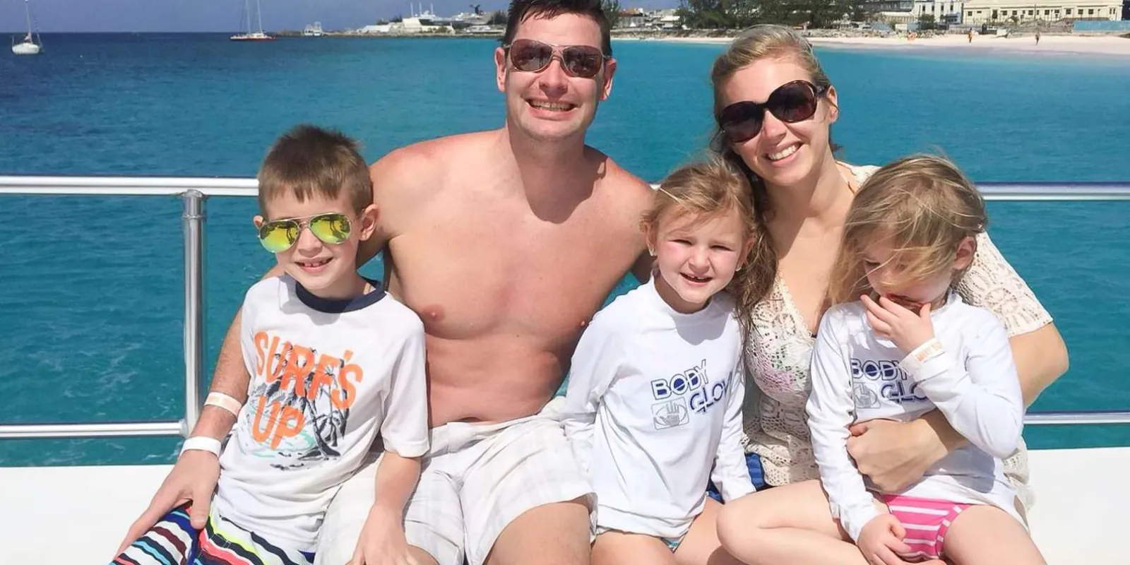 A family sitting on the boat's bow rail with Carlisle Bay and the beach behind them