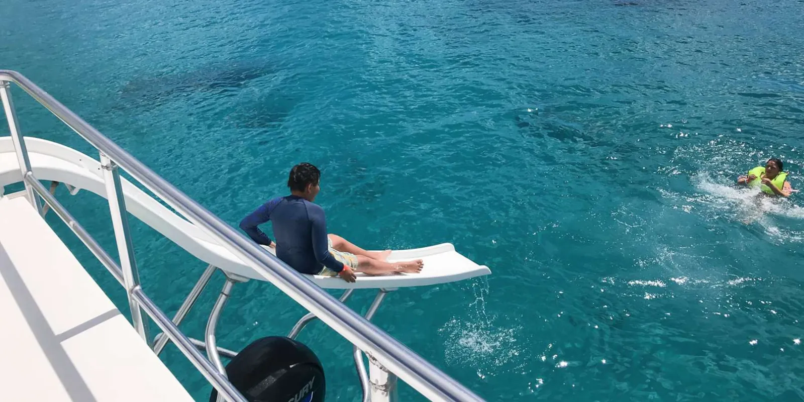 A swimmer sitting on the boat's bathing platform as another snorkeller swims back to the ladder
