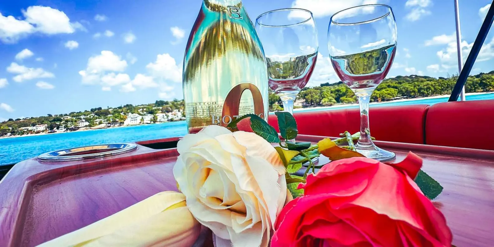 A bottle of sparkling wine and two glasses on the boat's table with the coastline behind