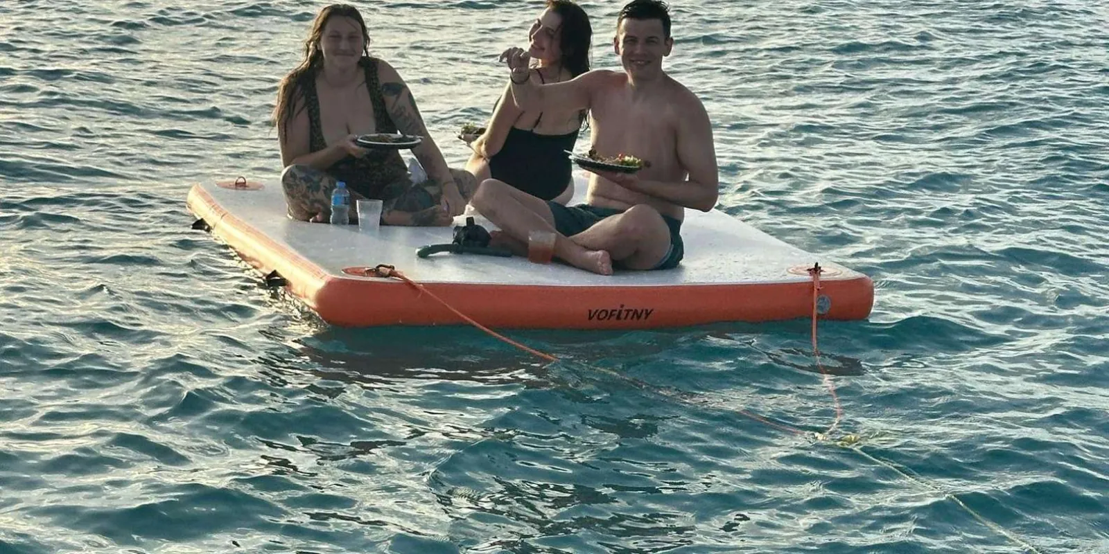Guests eating from plates on a floating platform moored beside the boat at dusk