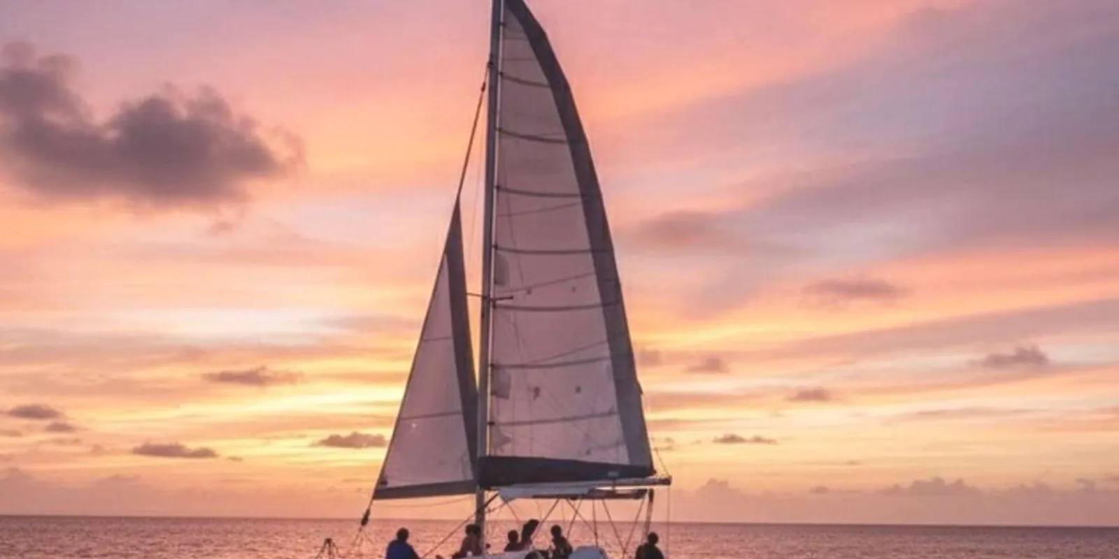 The charter catamaran sailing under a pink and gold Barbados sunset with guests on the foredeck